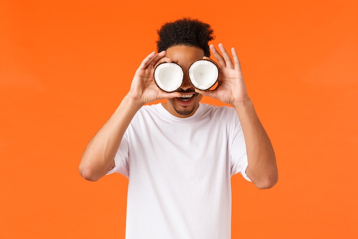 Free Photo excitement, holidays and vacation concept. amused and surprised african-american hipster guy with afro haircut, making glasses from coconuts on eyes, smiling wondered, standing orange background.