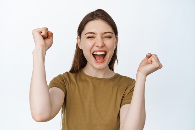 Excitement of win. Happy girl cheering and shouting from joy, celebrating victory with hands raised up and pleased face expression, triumphing on white.