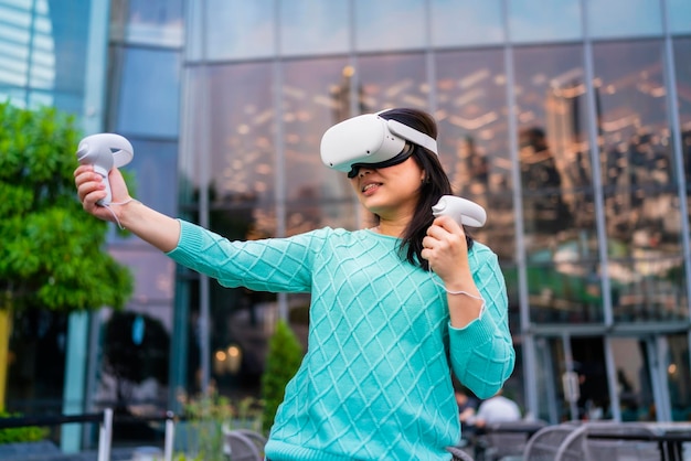 Exciting asian female gamer playing virtual reality augmented metaverse futuristic 3d gaming at outdoor of buildingBeautiful young woman playing game in virtual reality glasses with fun and joyful