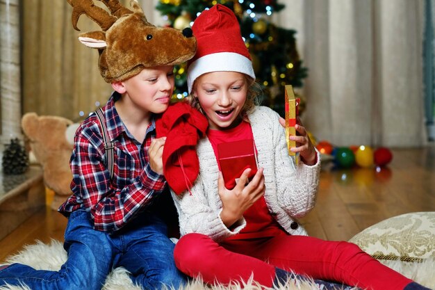 크리스마스 사슴 모자를 쓴 흥미진진한 귀여운 소년과 행복한 소녀는 크리스마스 장식된 방에 선물 상자를 들고 있습니다.