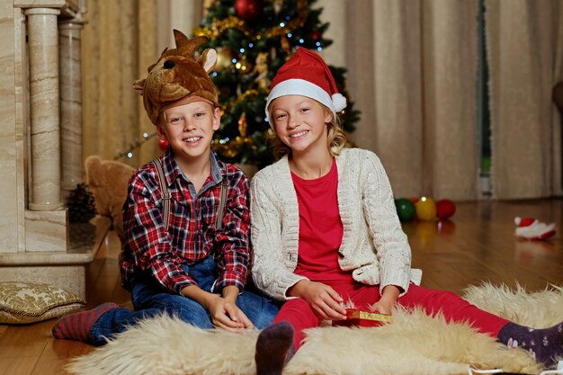 Exciting cute boy in Christmas deer's hat and happy girl holds gift box in Christmas decorated room.