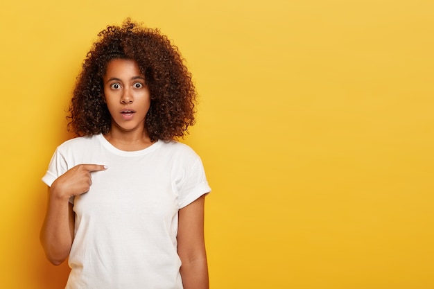 Excuse me? Indignant Afro American lady feels perplexed, points at herself, startled by offensive words, looks confusingly , wears white clothes, stands against yellow wall, blank space aside
