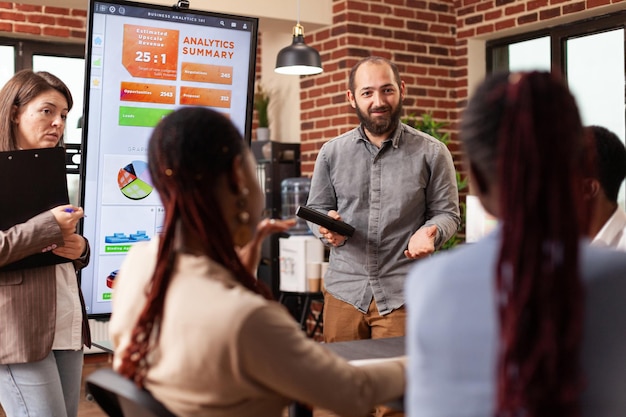 Foto gratuita direttore esecutivo in piedi accanto al monitor che mostra grafici di marketing che spiegano la strategia aziendale durante la riunione di lavoro nell'ufficio di avvio. diversi uomini d'affari che lavorano alla presentazione finanziaria