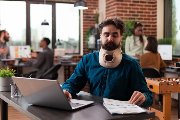 Executive manager with medical cervical neck collar while working at company turnover analyzing marketing strategy in brick wall startup office. Injured entrepreneur man planning business collaboratio