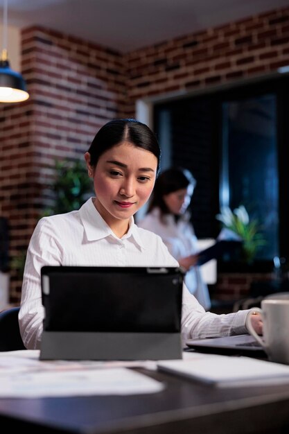 Executive manager working on electronic touchscreen device while developing marketing strategy. Asian businesswoman with modern tablet reviewing accounting documentation in office workspace.