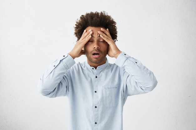 Free photo exhausted african male with bushy hair having headache standing with closed eyes