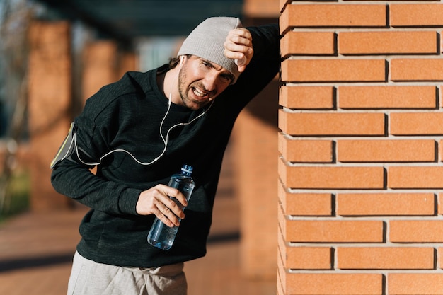 Uomo atletico esausto che si appoggia su un muro mentre fa una pausa d'acqua dopo l'esercizio all'aperto.