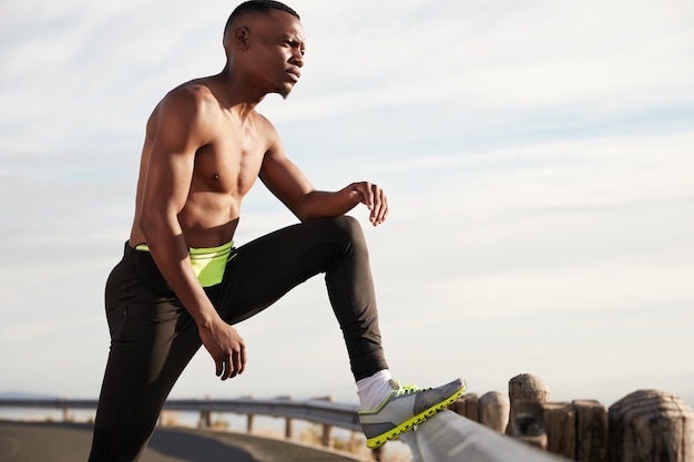 Exhausted black male runner rests after active run, wears sneakers, poses, thinks about future sport competitions, has hard sport workout. Jogging and motivation