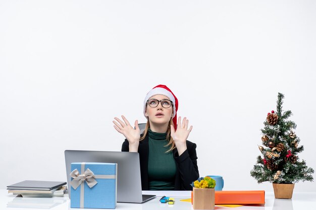 Exhausted business woman with santa claus hat sitting at a table with a Xsmas tree and a gift on it in the office on white background