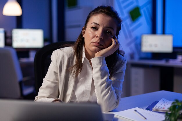 Изможденная бизнес-леди выглядит усталой в камеру, вздыхает, положив голову на ладонь поздно ночью в деловом офисе