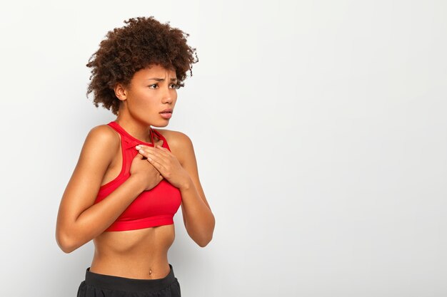 Exhausted curly female suffers from asthma breathing problems, keeps both hands on chest, wears red top