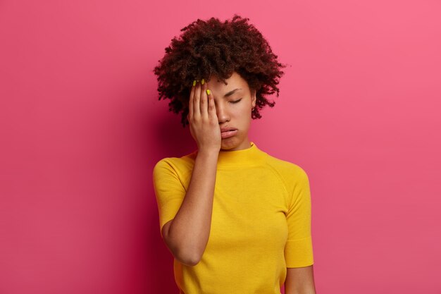 Измученная темнокожая молодая женщина прикрывает половину лица, вздыхает от усталости, имеет сонное выражение, закрывает глаза, носит желтую футболку, позирует на розовой стене. Женщина чувствует скуку и усталость