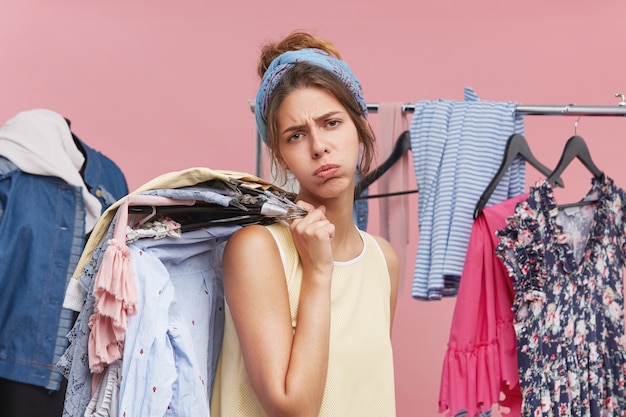 Esausto modello femminile che soffia le guance e che ha un'espressione stanca stando in piedi in camerino, tenendo grucce con vestiti sulla spalla, accigliato viso con insoddisfazione. stanchezza e shopping