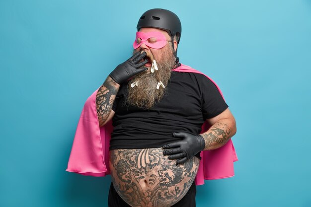 Exhausted male superhero yawns and covers mouth stands with big tattooed belly indoor being tired of doing different things models against blue wall