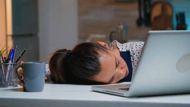 Exhausted overload business woman falling asleep on desk with open laptop monitor while working from home. Busy employee using modern technology network wireless doing overtime sleeping on table.