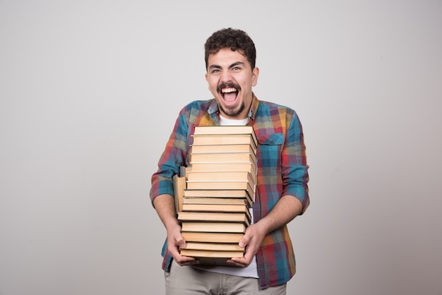 Exhausted student with pile of books screaming on gray background.