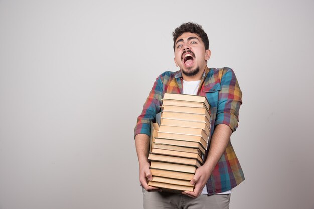 Exhausted student with pile of books screaming on gray background.