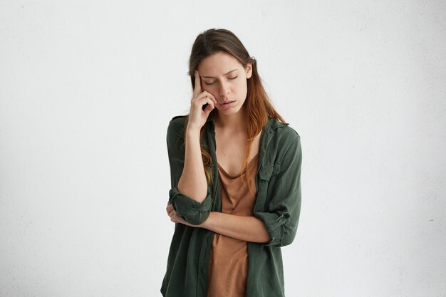 Exhausted and tired woman with dark hair standing against white wall with her eyes closed holding index finger on temple thinking about something. Upset female with tired expression.