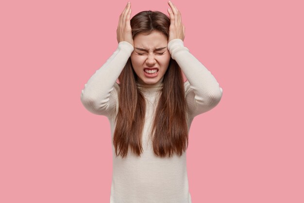 Exhausted worried brunette woman keeps hands on temples, clenches teeth, has headache or migraine, dressed in white outfit, isolated over pink background