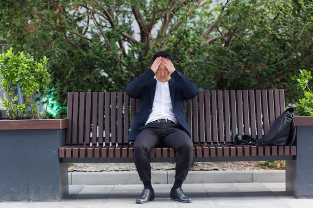 屋外​の​都市​公園​の​ベンチ​に​座っている​疲れ​果てた​若い​アジア人​ビジネスマン​の​サラリーマン​。​頭痛​で​過労​男性​を​強調し​、​頭​を​マッサージします​。​うつ病​の​疲れた​従業員​が​苦痛​の​病気​の​仕事​に​苦しんでいる
