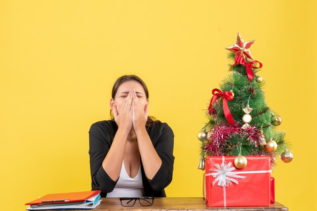 Exhausted young woman in suit near decorated Christmas tree at office on yellow 