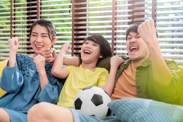 Exited Cheerful Joyful Asian parents And Son Watching Sports On TV Cheering Favorite Soccer Team Sitting Together On sofa couch At Home Weekend With Father Family time Online Tv Concept