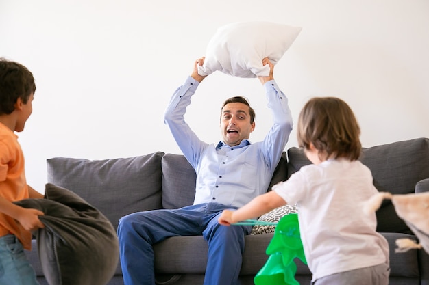 Exited dad sitting on couch and holding pillow above head. Happy children playing with father, fighting with pillows and having fun at home together. Childhood, holiday and game activity concept