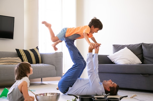 Exited father holding son on legs and lying on carpet. Cheerful Caucasian boys playing in living room with dad and utensils. Cute boy sitting on floor. Childhood, holiday and game activity concept