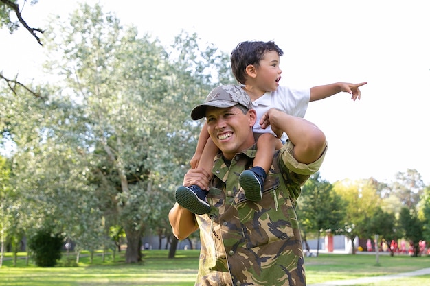 Exited little boy sitting on dad neck and pointing away. Caucasian father holding son legs, smiling, wearing army uniform and walking in park. Family reunion, fatherhood and returning home concept