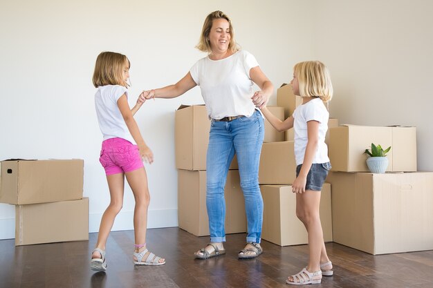 Exited mother standing and holding hands of two girls among unpacked boxes