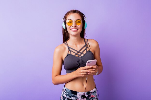 Exited sporty woman with  sincere  smiling posing in stylish active wear on studio. Using  smartphone   and listening   music.