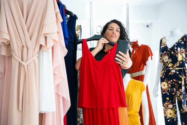Exited woman shopping in clothes store and consulting friend on cellphone, showing red dress on hanger. Medium shot. Boutique customer or communication concept