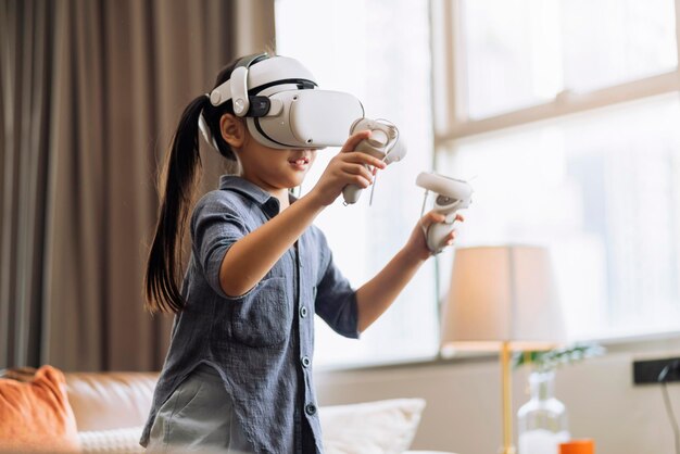 Exiting asian child female enjoy metaverse gaming with wearable vr headset with control handle playing sport gaming online in living room at homehome technology female daughter using vr technology