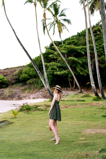 Exotic summer portrait of pretty blonde woman posing at exotic island, wearing dress and straw hat. Luxury vacation on island.