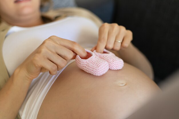 妊娠中の母親はピンクのベビーシューズを大きな裸の腹に適用します。
