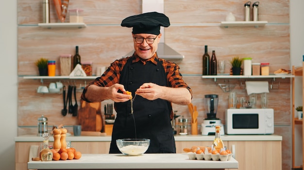 Experienced baker man craking eggs for baking wearing apron enjoying hobby. Retired elderly chef with bonete mixing by hand, kneading in glass bowl pastry ingredients preparing homemade cake