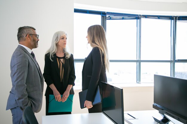 Experienced colleagues standing in office room and looking at each other. Professional content CEO and pretty businesswomen discussing work project. Business, communication and corporation concept