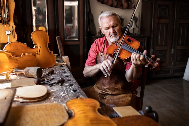 Опытный седой старший столяр в кожаном фартуке сидит в своей столярной мастерской и играет на скрипке