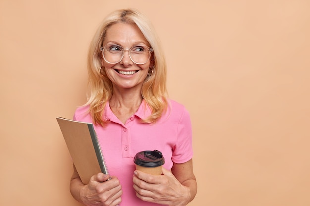 Foto gratuita tutor femminile intelligente esperta dà lezioni private bevande caffè da asporto tiene taccuini sorrisi indossa piacevolmente occhiali ottici maglietta rosa isolata sopra lo spazio della copia muro marrone