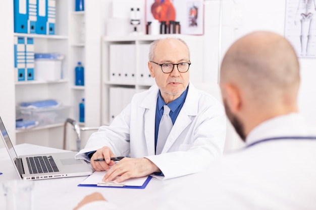 Experienced mature doctor explaining patient diagnosis to young medical physician during conference. Clinic expert therapist talking with colleagues about disease, medicine professional.