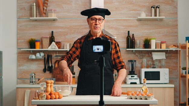 Experienced senior chef recording tutorial with food preparation in kitchen. Retired blogger influencer using internet technology communicating shooting blogging on social media with digital equipment