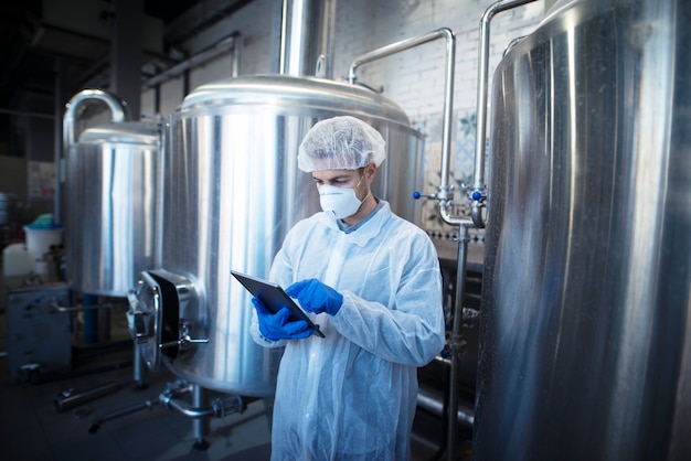 Опытный технолог в белой защитной униформе держит таблетку и контролирует производство продуктов питания на перерабатывающем заводе.