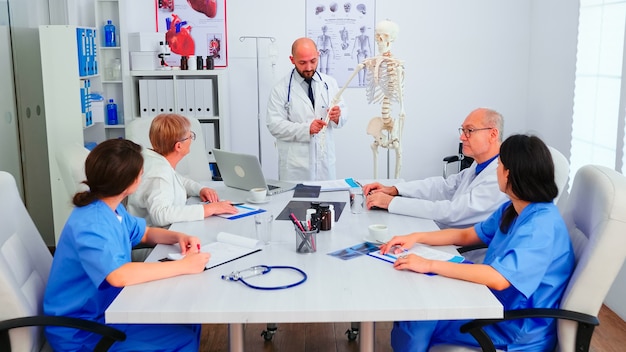 인간 골격 모델을 사용하여 회의실에서 의료진을 위한 의료 세미나 중에 방사선 사진을 들고 있는 전문 의사. 질병에 대해 동료와 이야기하는 클리닉 치료사, 의학 전문가