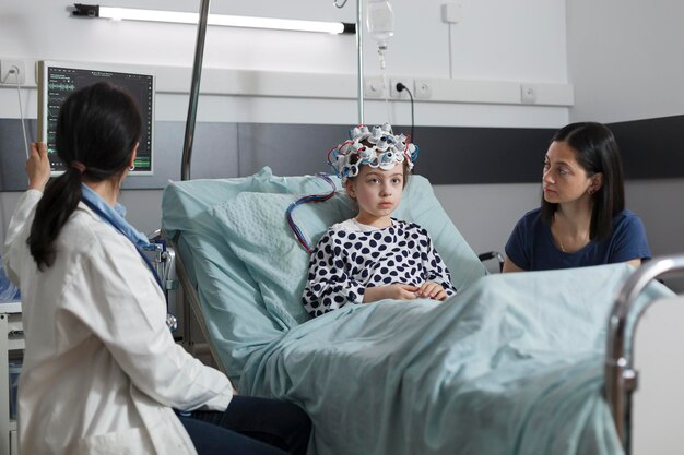 Expert pediatrician monitoring hospitalized girl brain condition using EEG headset. Neurology specialist measuring neurologic disease evolution of sick kid sitting in pediatric healthcare facility.