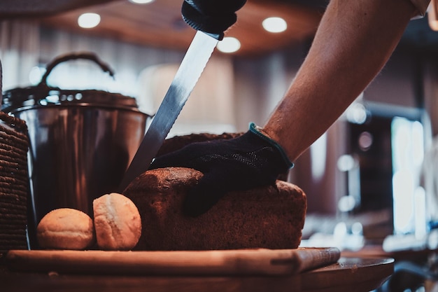 Expirienced baker in protective gloves is slicing bread for daily breakfast at restaurant.