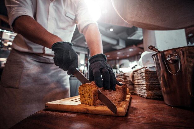 Expirienced baker in protective gloves is slicing bread for daily breakfast at restaurant.