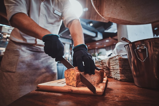 Expirienced baker in protective gloves is slicing bread for daily breakfast at restaurant.