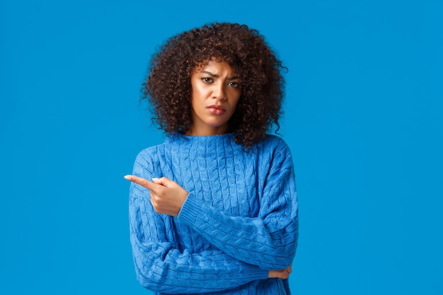 Объясните мне, что это. недовольная мрачная и неудовлетворенная афро-американская девушка с афро-стрижкой, указывая пальцем влево, скептически и неодобрительно хмурясь, синий фон