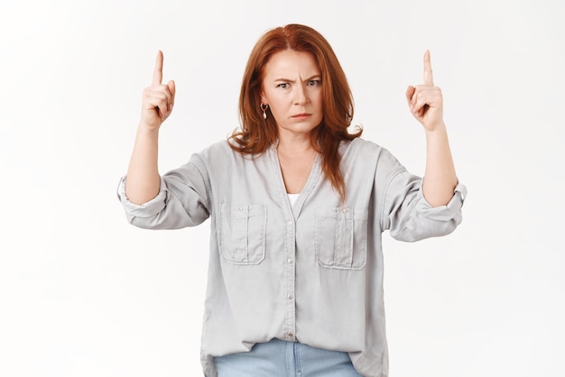 Explain yourself young man Angry displeased middleaged redhead mother frowning pointing up index finger dangerous annoyed stare camera scolding child bad behaviour standing white background upset