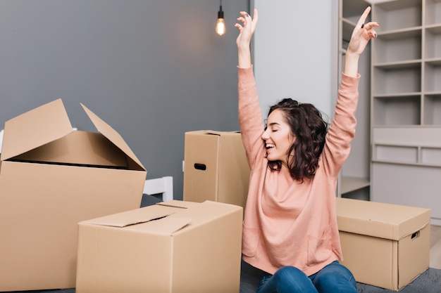 Expressing brightful true emotions, positivity of young pretty woman with short brunette curly hair on bed surround carton boxes in modern apartment. Enjoying moving, happiness at new home